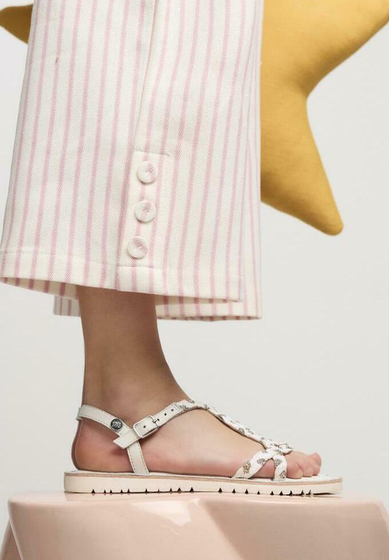 Sandalias blancas con suela plana, correas decorativas y cierre de hebilla. Combinadas con pantalones rayados en rosa claro con detalles de botones.