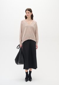 Beige Strickpullover mit V-Ausschnitt, kombiniert mit einem schwarzen knöchellangen Rock, schwarzen Socken und spitzen Schuhen, und hält eine schwarze Tasche.