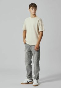 Beige t-shirt med korta ärmar, grå jeans och vita sneakers med svarta detaljer. Modellen står med händerna i fickorna mot en neutral bakgrund.