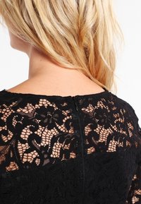 Černé krajkové šaty s květinovými vzory, průhledné rukávy a viditelný zip na zadní straně. Texturovaný materiál a těsně přiléhající střih.