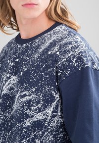 Sudadera de marina con patrón de salpicaduras blancas, hecha de algodón. Presenta un cuello redondo y mangas lisas en un tono de marina contrastante.