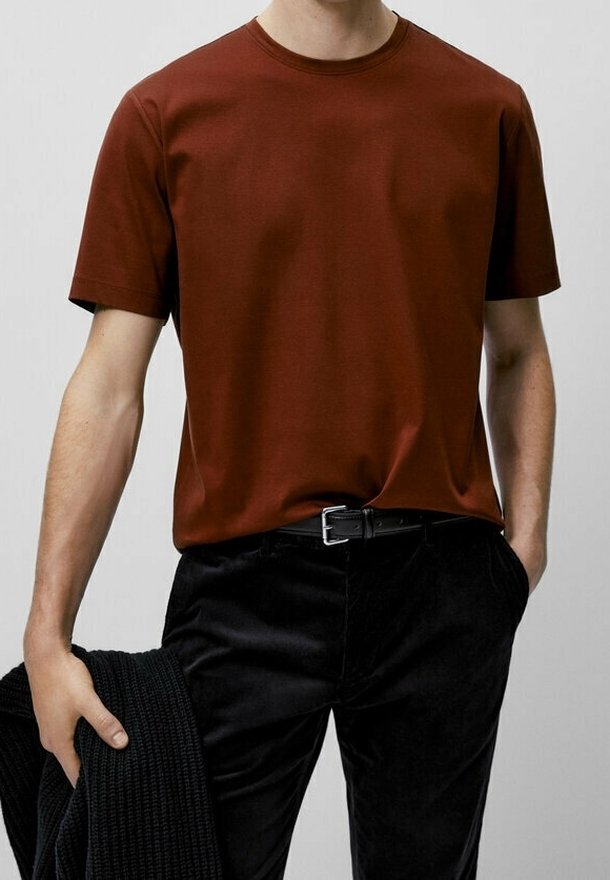 Personne portant une chemise à manches courtes couleur rouille et un pantalon noir, tenant une veste noire texturée dans une main.