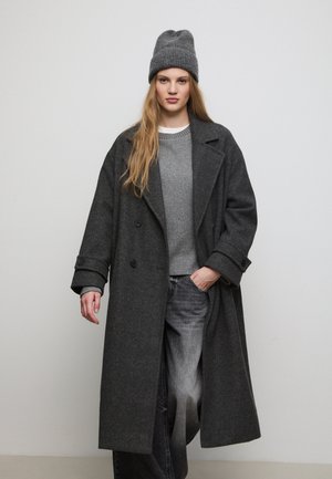 Cappotto di lana grigio con ampi risvolti, doppi bottoni e tasche laterali, indossato sopra un maglione grigio e jeans grigi sbiaditi. Berretto di maglia grigio.