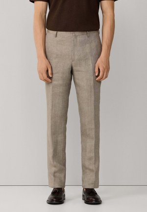 Hombre con pantalones de sastre color marrón claro, mocasines de cuero marrón oscuro con borlas y camisa de manga corta marrón oscuro, sobre un fondo liso.