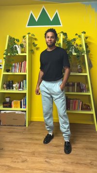 Μαύρο πουκάμισο με κοντά μανίκια, ανοιχτό μπλε παντελόνι φόρμας με ελαστικά μανσέτες και μαύρα αθλητικά παπούτσια. Το φόντο περιλαμβάνει κίτρινους τοίχους και ράφια με πράσινα φυτά.