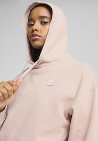Sudadera rosa claro con un bolsillo frontal, capucha con cordón y un pequeño parche bordado con el logo. Textura de tela suave. El modelo sostiene el dobladillo.
