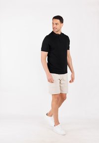 Polo shirt nero a maniche corte con colletto e chiusura a bottoni, abbinato a pantaloni beige e sneakers bianche, in piedi davanti a uno sfondo semplice.