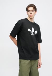 Čierne tričko s krátkym rukávom vyrobené z bavlny s veľkým bielym logom Adidas a tromi prúžkami, nosené s zelenými cargo šortkami.