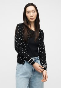 Γυναίκα που ρυθμίζει το μανίκι ενός μαύρου cardigan με λευκές πουά, φοράει μαύρο τοπ και ανοιχτόχρωμα τζιν ψηλόμεσα, μπροστά σε απλό φόντο.