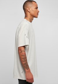 Lys grå oversized t-skjorte laget av myk bomull, med rå kanter og slitte detaljer på ermene. Sideprofilbilde.