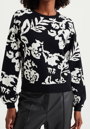 Pull noir avec de grands motifs floraux blancs, porté avec un pantalon noir taille haute sur une personne aux cheveux bouclés.