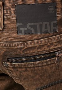 Pantalones vaqueros de mezclilla marrón con un parche de cuero negro con la marca "G-STAR", costuras visibles y un bolsillo delantero con cremallera.