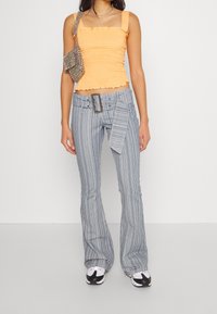 Oranges, gerafftes Tanktop mit Rüschensaum, kombiniert mit blauen, vertikal gestreiften, ausgestellten Hosen mit großer Gürtelschnalle und asymmetrischem Gürtel-Detail.