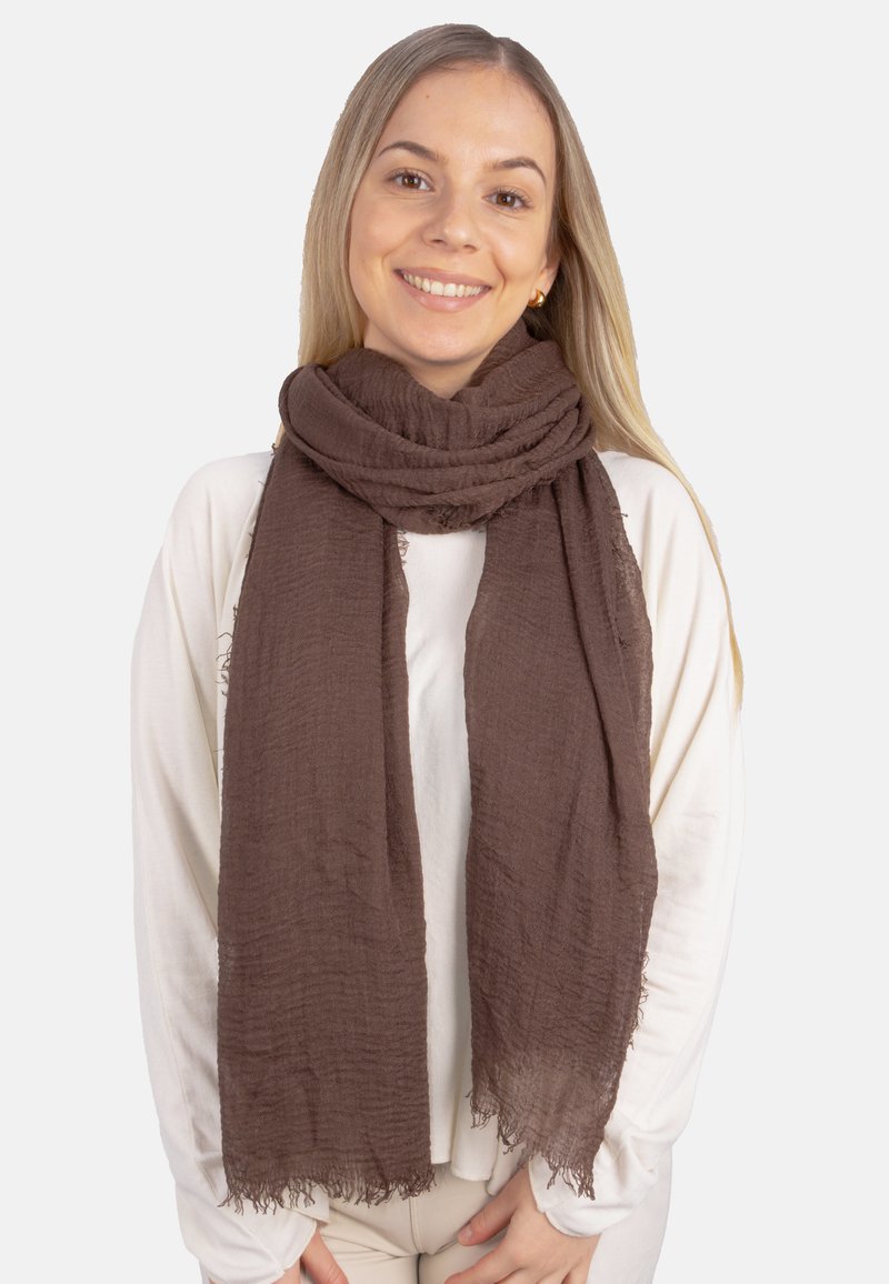 Écharpe marron texturée avec des bords effilochés, enroulée autour du cou, portée par-dessus un haut de couleur claire. Matériau doux et léger.