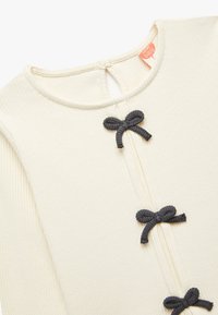 Crème ribbed top met drie decoratieve donkergrijze strikken langs de voorkant, met een ronde halslijn en een kleine opening met sleuteltje aan de achterkant.