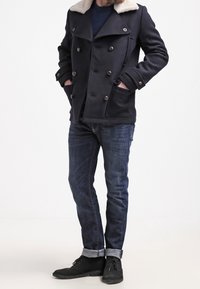 Marintröja med dubbelknäppt design, knappdetaljer och krämfärgad fuskpälskrage, bärs över en marinblå skjorta och mörka jeans med uppvikta fållar.