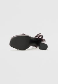 Glossy burgundy high-heeled shoe with a textured black sole, featuring a strap design and a rounded toe.