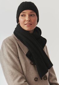 Gorro de lana negro y bufanda a juego con textura acanalada, usados sobre un abrigo gris claro con grandes botones marrones.