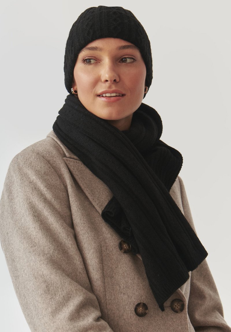 Gorro de lana negro y bufanda a juego con textura acanalada, usados sobre un abrigo gris claro con grandes botones marrones.