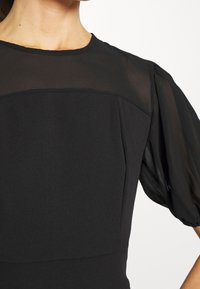 Detailní záběr ženy v černých šatech s průhledným živůtkem a krátkými nabíranými rukávy na jednobodovém pozadí.
