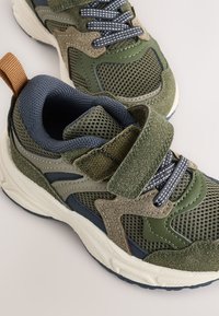 Žalios ir tamsiai mėlynos sportinės bateliai su tinklinio tipo viršumi, zomšos akcentais ir lipniu dirželiu. Su minkštinimu ir tekstūruotu guminio pado paviršiumi.