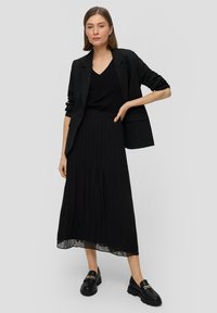 Zwarte geplooide rok met een doorzichtige zoom, gecombineerd met een zwarte v-hals top en een op maat gemaakt blazer. Het schoeisel heeft een chunky ontwerp met metalen accenten.