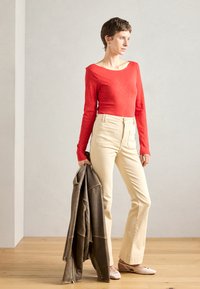 Piros, hosszú ujjú felső, krém színű, bővülő nadrág és világos rózsaszín balerina cipő. Egy barna bőrdzsekit tart a kezében. Simaság és testhez álló szabás jellemzi.