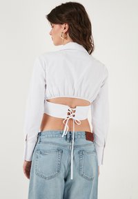 Camicia bianca corta con maniche lunghe, dettaglio in pizzo sulla schiena e design aperto sulla schiena. Abbinata a jeans high-waisted azzurri chiari.