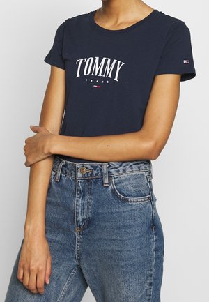Person trägt ein dunkelblaues Tommy Jeans T-Shirt und hoch taillierte blaue Denim-Jeans, die Arme vor der Taille verschränkt.