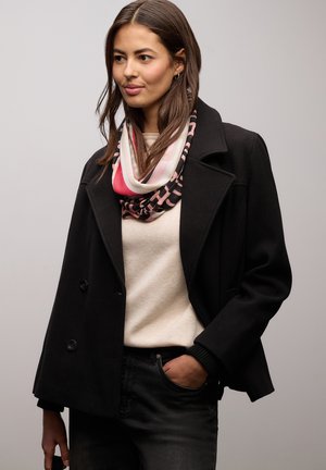 Manteau noir croisé à double boutonnage avec de grands revers, porté sur un pull crème. Une écharpe infinie à motifs rose et noir est drapée autour du cou.