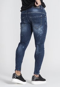 Jeans de mezclilla azul delgado con un lavado ligeramente descolorido, detalles en el bolsillo trasero y sutiles salpicaduras de pintura. Combinados con zapatillas negras con estampado.