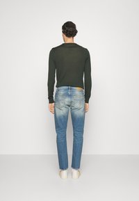 Camicia a maniche lunghe verde scuro abbinata a jeans in denim chiaro e sbiadito. I jeans presentano un design a cinque tasche con una patch in pelle beige.