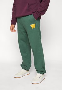Gröna sweatshirtbyxor med elastisk midja, muddade anklar och en gulbroderad "W"-logotyp. Matchas med vita sneakers.