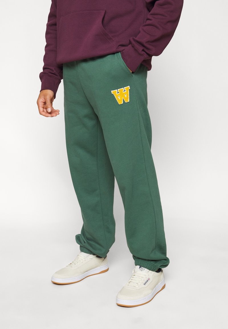 Gröna sweatshirtbyxor med elastisk midja, muddade anklar och en gulbroderad "W"-logotyp. Matchas med vita sneakers.