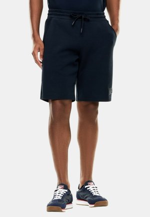 Persona che indossa pantaloncini blu navy con coulisse e sneakers blu navy con lacci bianchi, in piedi con le mani nelle tasche davanti a uno sfondo semplice.