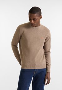 Beiger Strickpullover mit geripptem Rundhalsausschnitt und Bündchen. Rot gesticktes Logo auf der linken Brust. Getragen mit dunkelblauen Jeans.