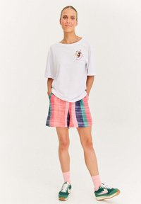 Žena stojící s rukama v kapsách, oblečená v bílé oversized tričku s grafikou, multicolor proužkovaných šortkách, růžových ponožkách a zelených teniskách.