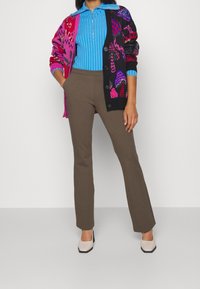 Bunt gemusterter Cardigan in Pink, Schwarz und Lila, über einem hellblauen Zip-Up-Top getragen, kombiniert mit braunen Schlaghosen und hellen Schuhen.