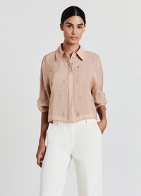 Blusa rosa pallido a maniche corte in tessuto leggero con colletto a bottoni, caratterizzata da delicati ricami e un fit rilassato, abbinata a pantaloni bianchi.