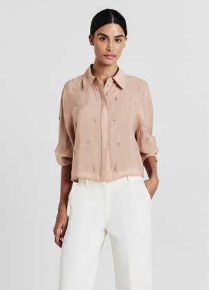 Blush-pink Bluse mit kurzen Ärmeln aus leichtem Stoff und Knopfkragen, verziert mit zarter Stickerei und in lockerer Passform, kombiniert mit weißen Hosen.