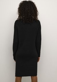 Robe en maille noire à manches longues, ourlet côtelé, coupe décontractée, vue de dos sur une personne aux cheveux bouclés.