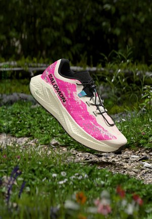 Lett rosa og beige Salomon løpesko i luften over en gresskledd, steinete sti med skog i bakgrunnen.