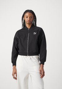 Veste bomber noire courte avec fermeture éclair sur le devant, poignets côtelés et un logo Puma blanc sur la poitrine. Associée à un pantalon large blanc.