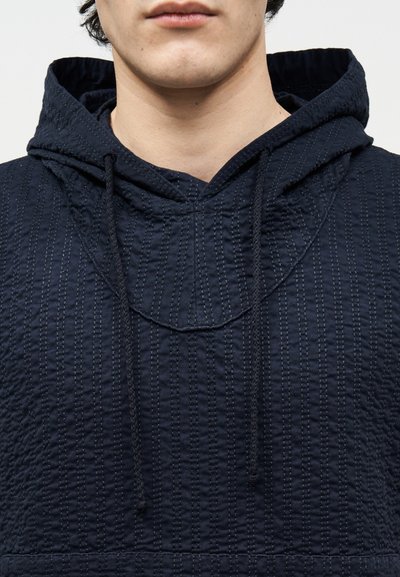 Jongere draagt een marineblauwe, gestructureerde hoodie met zichtbare trekkoorden, getoond van kin tot midden torso tegen een neutrale achtergrond.