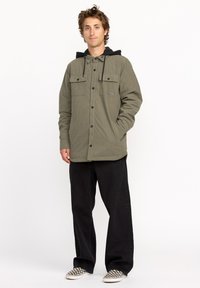 Veste boutonnée olive avec capuche noire, deux poches poitrine et un tissu texturé. Associée à un pantalon noir et des chaussures à enfiler à carreaux.