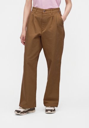 Persona che indossa pantaloni marroni larghi con pieghe, mani nelle tasche, abbinati a sneaker bianche e marroni e una maglietta rosa chiaro.