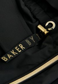 Črn material z gladko teksturo ima zlat zadrgo in črno oznako z napisom "BAKER BY T" v zlatem napisu.