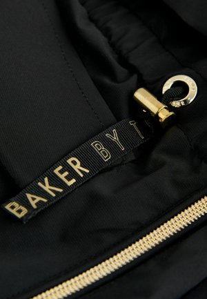 Schwarzer Stoff mit glatter Textur, versehen mit einem goldenen Reißverschluss und einem schwarzen Etikett mit "BAKER BY T" in goldener Schrift.