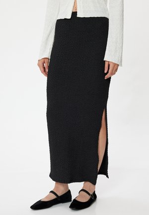 Zwarte, textuurachtige maxi rok met een zijsplit, gecombineerd met zwarte platte schoenen. De stof heeft een gekreukeld uiterlijk en een getailleerde pasvorm.