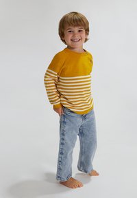 Gestreifter gelb-weißer Langarmpullover mit Knöpfen, kombiniert mit hellblauen Jeans. Kind steht barfuß und lächelt.